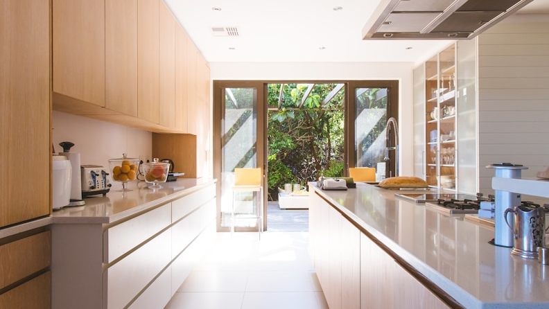 A galley kitchen with pale oak cabinetry and white walls opening onto a garden